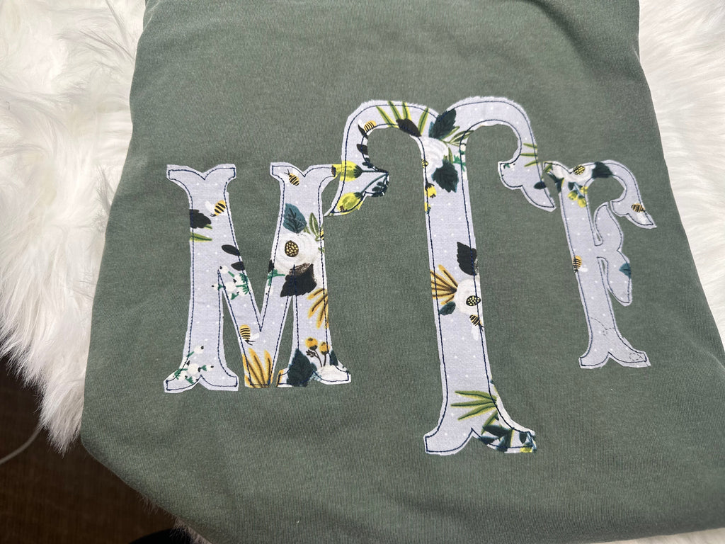 Botanical floral MTR serif monogram appliqué on a sage shirt, shown upright