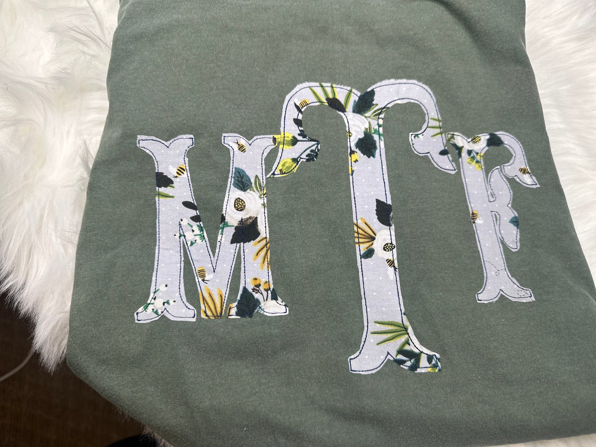 Botanical floral MTR serif monogram appliqué on a sage shirt, shown upright